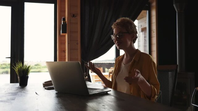 A smiling caucasian woman with short hair and glasses is engaged in a video call on her laptop while sitting at a wooden table in a cozy cabin