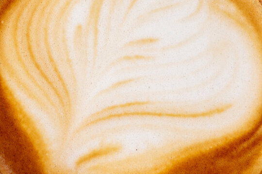 A coffee background made of close up latte art foam shows delicate feathered patterns in warm caramel and cream tones filling the entire frame with no cup edges visible