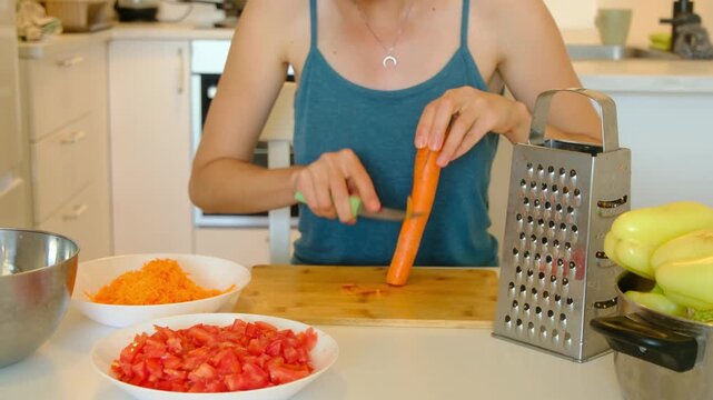 woman preparing ingredients for traditional stuffed peppers dish. White plate filled with chopped red tomatoes stands near yellow bell peppers and onions.