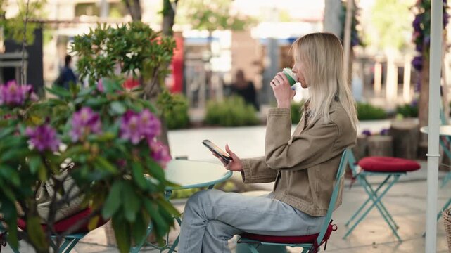 Young blonde woman sitting at an outdoor cafe table, leisurely drinking coffee from a paper cup and holding a smartphone while enjoying a quiet moment on a city street in slow motion