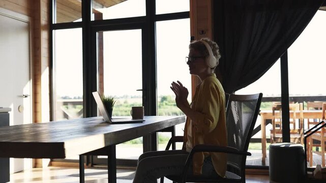 A young woman wearing headphones and glasses is engaged in a video call on her laptop while sitting at a wooden desk in a bright, modern room with large windows