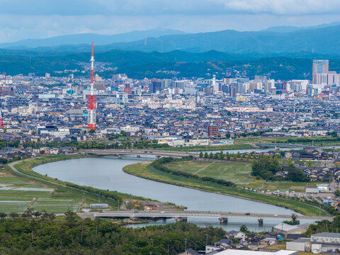 Aerial view of the Sai River winding through the urban landscape with a prominent red and white television tower and distant mountains, Kanazawa, Ishikawa, Japan.