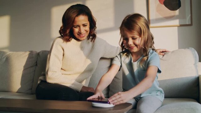 Smiling mother in sweater guiding daughter placing purple phone on wireless puck at home to charge