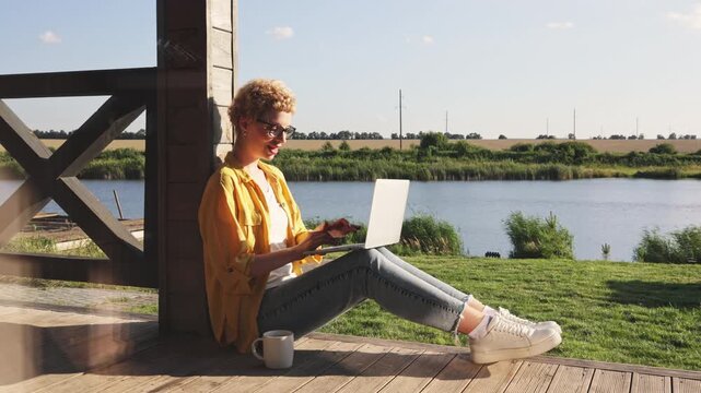 A happy caucasian woman enjoys a sunny day while working on her laptop by a tranquil lake, embracing a remote work lifestyle