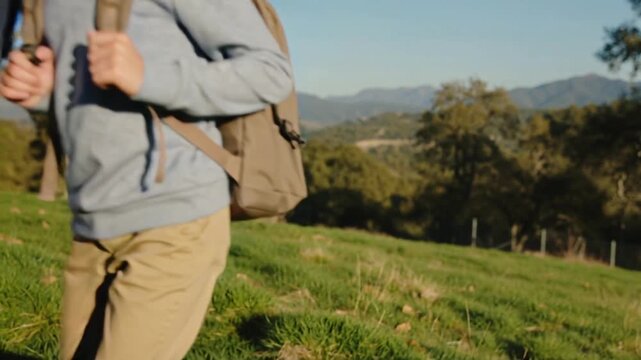 Reaching out, dad in navy hoodie tightening boy's tan backpack straps on grassy hill, starting walk