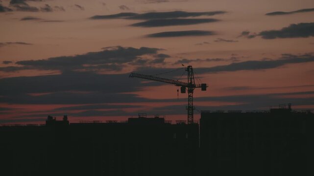 Dramatic timelapse showing a construction crane silhouette working against a beautiful evening sky with colorful clouds moving over the dark cityscape at dusk, highlighting urban development