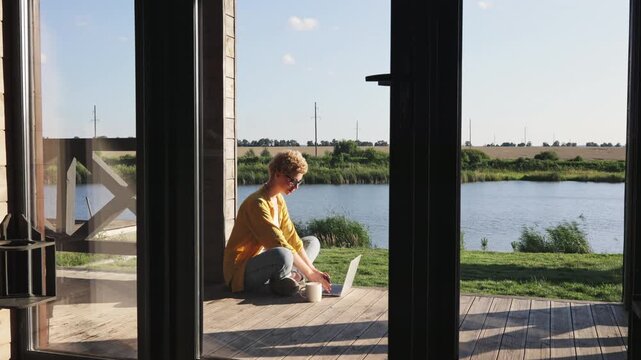 A caucasian woman in casual clothes sits in a meditative pose on a wooden deck overlooking a tranquil lake and fields