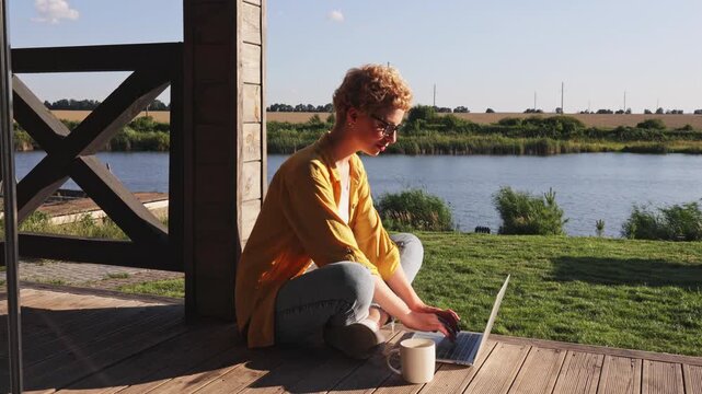 A caucasian woman in glasses sits cross-legged on a wooden deck, meditating with a laptop and mug nearby, enjoying a serene lakeside view