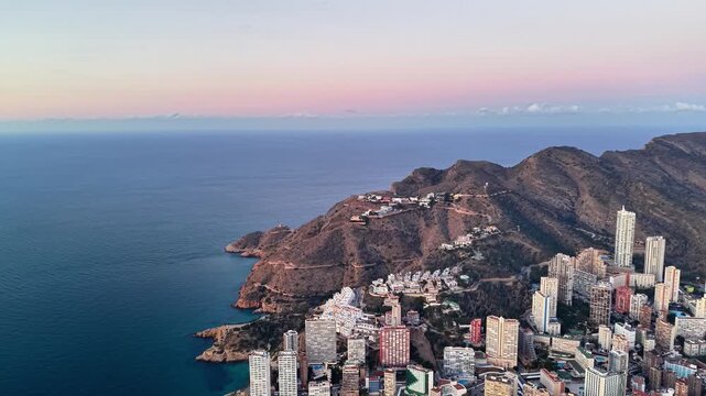 Aerial coastal skyline at sunrise, soft pastel sky and calm ocean, highrise buildings clustered on rocky