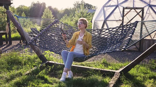 A caucasian woman with short curly hair and glasses is relaxing in a hammock while recording a video on her smartphone with a selfie stick