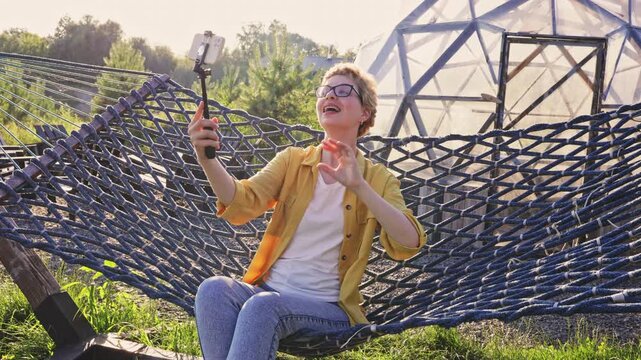 A happy young woman with short blonde hair and glasses is relaxing in a hammock and taking a selfie with her smartphone on a selfie stick