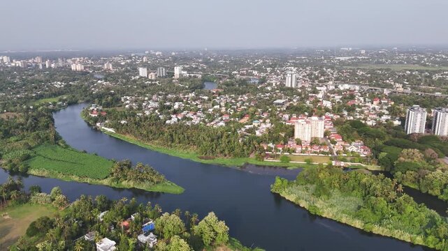 Drone shot capturing the scenic riverfront of Aluva, where the winding Periyar River separates dense greenery from vibrant residential communities and modern high-rise buildings in the distance.
