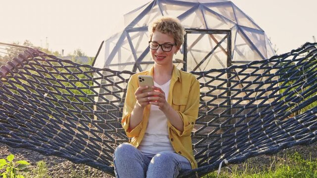 A young caucasian woman wearing glasses and a yellow shirt sits in a hammock outdoors, looking at her smartphone with a smile