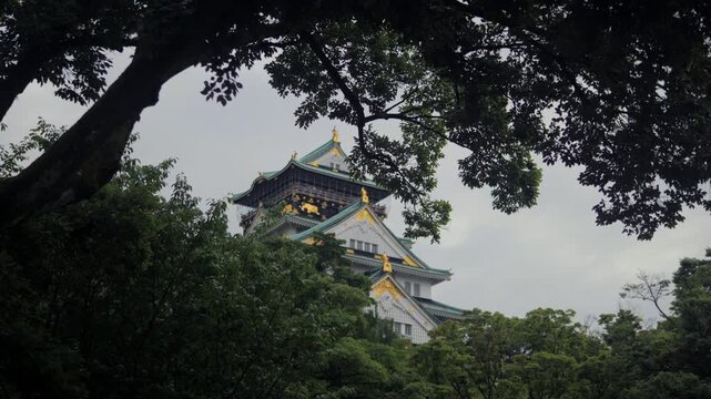 Traveling frontal shot of Osaka Castle emerging through lush green trees under cloudy skies.