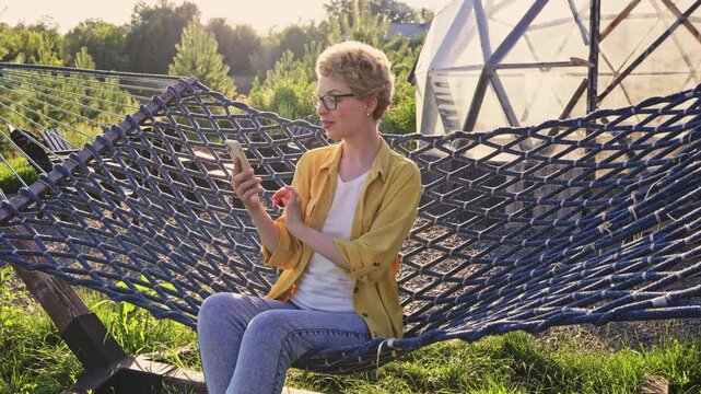 A caucasian woman wearing glasses and a yellow shirt is relaxing in a hammock while holding her phone and engaging in a video call outdoors