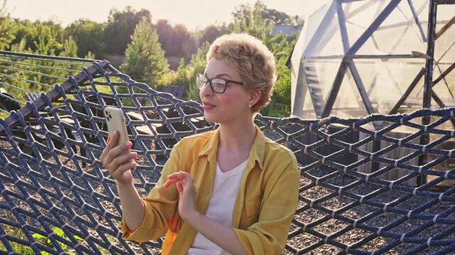 A smiling caucasian woman with short blonde hair and glasses is relaxing in a hammock while holding her phone and waving during a video call outdoors