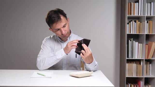 Man examining an empty wallet, reflecting on financial difficulties and budgeting challenges with a calculator and notebook on a white desk beside a bookshelf