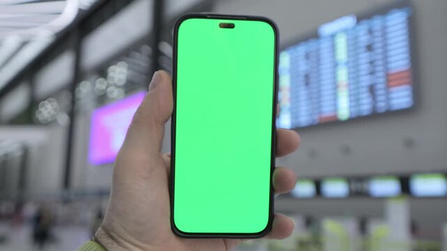 Hand holds smartphone with green screen. Background shows airport terminal with flight boards. Focus on phone, blurred surroundings. Realistic indoor lighting. No animals or plants visible