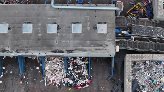 Plastic waste enters the facility for sorting and processing. Large piles of recyclable materials are visible outside. Workers process waste for resource recovery in an industrial environment.