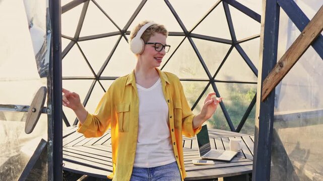A young Caucasian woman with short blonde hair and glasses enjoys music and dances inside gazebo, with a laptop on a nearby table