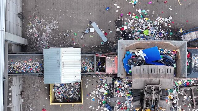 Plastic waste moves through a sorting system at a recycling facility. Various types of plastic are separated for processing. The area shows piles of waste being collected and sorted.