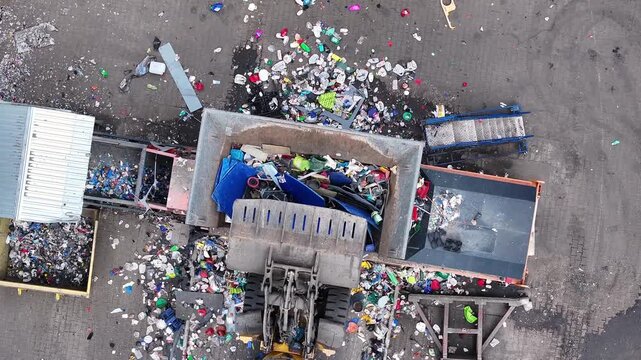 A large machine processes mixed plastic waste at an industrial recycling plant. Workers sort materials for recovery and recycling. The area is busy with trucks delivering waste.