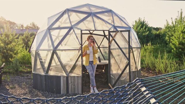 A cheerful Caucasian woman talks on her phone, holding a cup, standing relaxed by the entrance of a modern geodesic dome greenhouse gazebo, with a hammock in foreground.