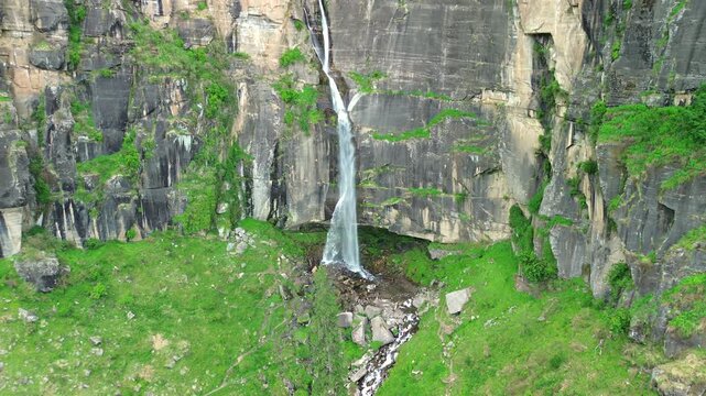 Drone slow pull-out shot revealing a majestic narrow waterfall plunging down a high, vertical black stone cliff surrounded by green grass patches in Manali, Himachal Pradesh, India