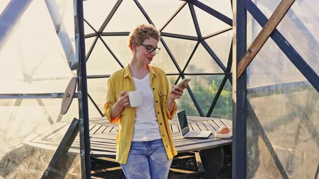 Attractive Caucasian woman holds a coffee mug and phone, smile directly at camera. She stands inside a clear, modern gazebo with a laptop on a wooden table, evoke a relaxed and productive atmosphere.