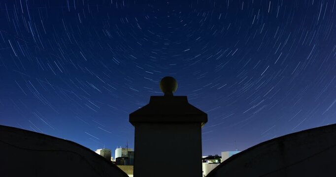 Timelapse of stacked star trails forming circular patterns above rooftops and chimney silhouettes in Alcantarilla, Murcia, Spain.