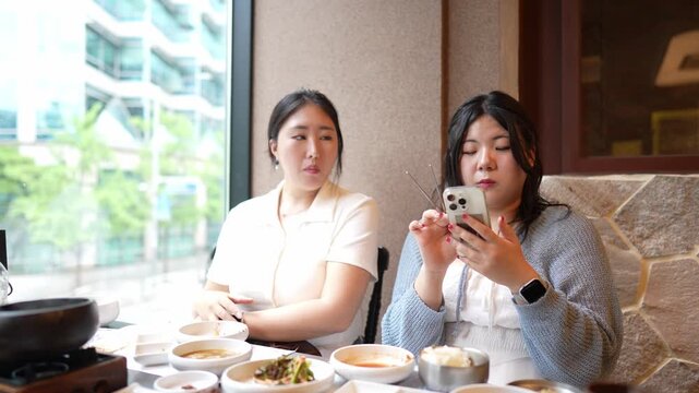 Two women enjoy a meal at a cozy restaurant. One sips soup while the other scrolls through her phone. They sit near a window with city views outside. Their expressions are calm, focused, and relaxed