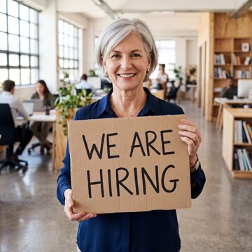 Smiling mature caucasian businesswoman holding a we are hiring cardboard sign in a busy modern office