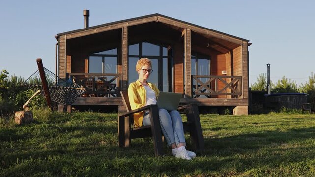 A caucasian woman with short blonde hair wearing glasses and a yellow shirt sits in a chair with a laptop in front of a wooden cabin