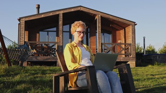 A young woman with short blonde hair and glasses sits in a chair with a laptop on her lap, enjoying the sunshine in front of a wooden cabin