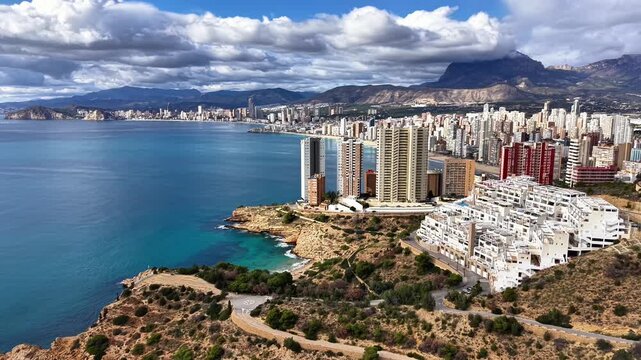Aerial panoramic skyline over turquoise bay showcasing white terraced villas, highrise towers, rugged cliffs