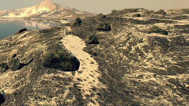 A rocky area with visible patterns leads down to a body of water. Mountains rise in the distance under a clear sky. The scene is bright and shows signs of movement in the sand.