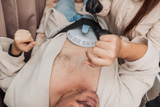 Middle-aged Hispanic man undergoing Emsculpt HIFEM body contouring treatment as an esthetician secures the abdominal applicator for non-invasive muscle strengthening and body sculpting.