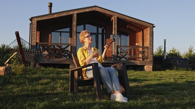 A caucasian woman with short blonde hair sits in a chair on the grass in front of a wooden cabin, recording a video with her smartphone on a gimbal