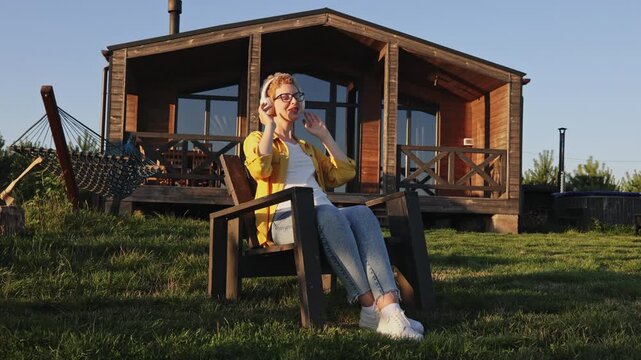 A young woman with headphones on enjoys music while relaxing in a chair outside her wooden cabin on a sunny day