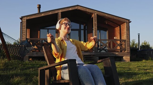 A young woman with short blonde hair and glasses sits in a chair outside a wooden cabin, enjoying music through her headphones