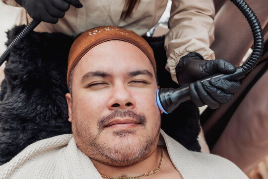 Professional esthetician performing a Hydra Oxygen Facial with a facial ultrasound sonophoresis probe on cheek of a middle-aged man to enhance serum absorption and skin hydration.