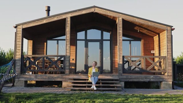 A caucasian woman wearing glasses and a yellow jacket sits on the steps of a wooden cabin with a laptop on her lap