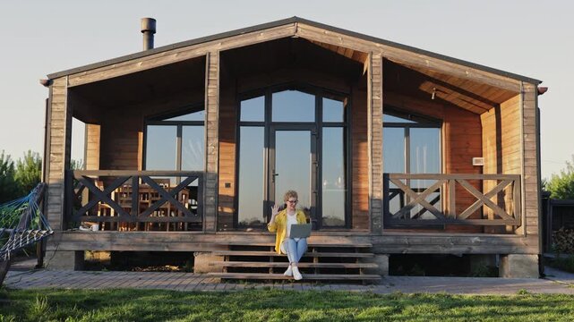 A young woman with curly hair sits on the steps of a wooden cabin, waving at her laptop screen while working remotely in a natural setting