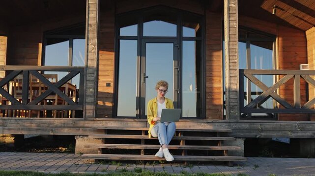 A young caucasian woman with short curly hair and glasses sits on wooden steps outside a cabin, working on her laptop and looking up thoughtfully