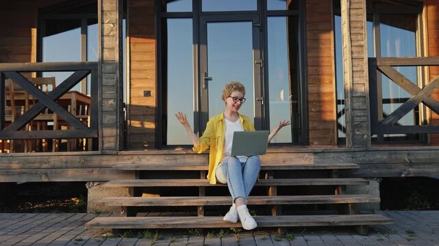 A young woman with curly hair and glasses sits on wooden steps outside a modern cabin, engaged in a video call on her laptop