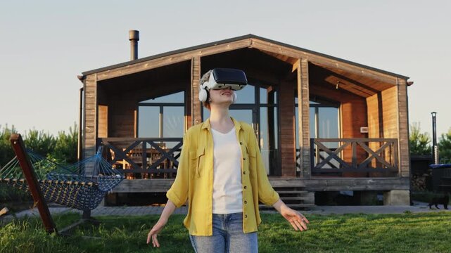 A caucasian woman wearing a VR headset and headphones stands in front of a wooden house, gesturing with her hands as she experiences virtual reality