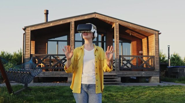 A caucasian woman wearing a VR headset and headphones is interacting with a virtual world while standing in front of a wooden house