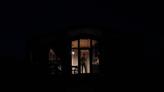 A solitary woman stands in a warmly lit room at night, silhouetted against the darkness outside