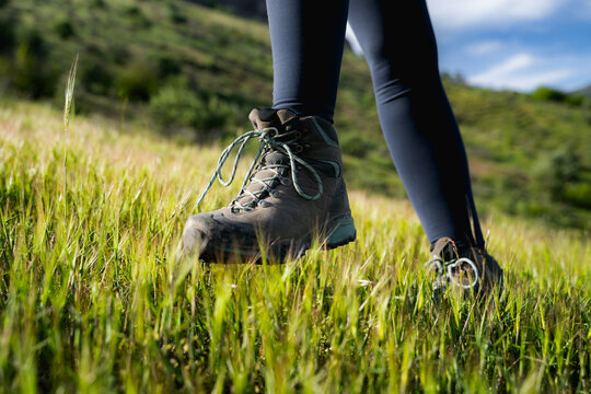 Close-up of a woman&rsquo;s trekking boots stepping through green grass on a mountain trail during a summer hike.