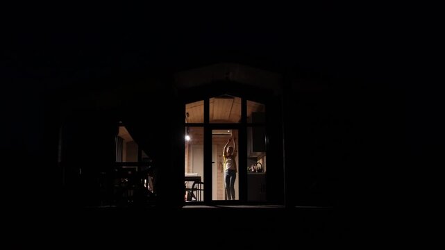 A young woman joyfully dances with her arms raised in a brightly lit room at night, seen through a large glass door